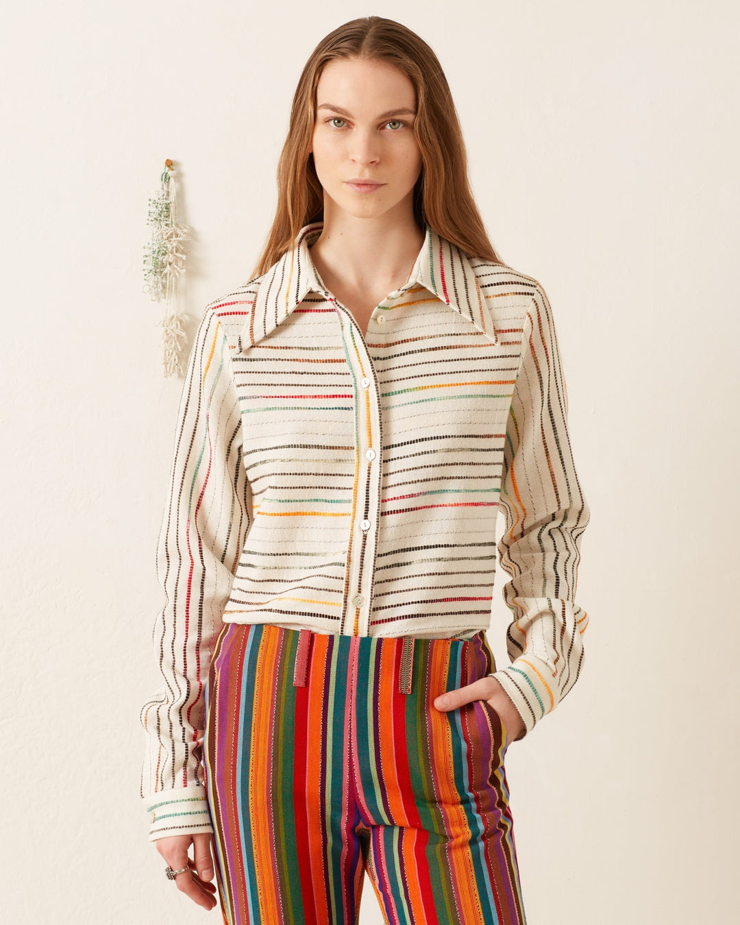 A person wearing the Dolly Chroma Weave Shirt, featuring a vintage-inspired '70s-style wing collar, paired with multicolored striped pants, stands against a beige background, highlighting the unique texture of hand-loomed cotton.