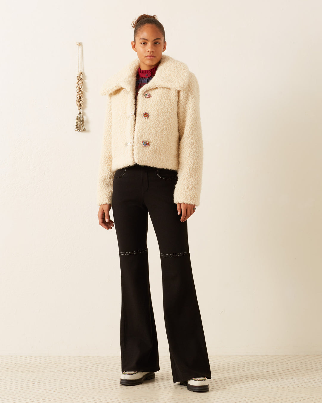 A person wearing a Colby Suri Wool Jacket with an oversized collar and black flared pants stands on a light-colored floor. The plain wall behind them features a decorative hanging. They have a neutral expression, and their hair is tied up.