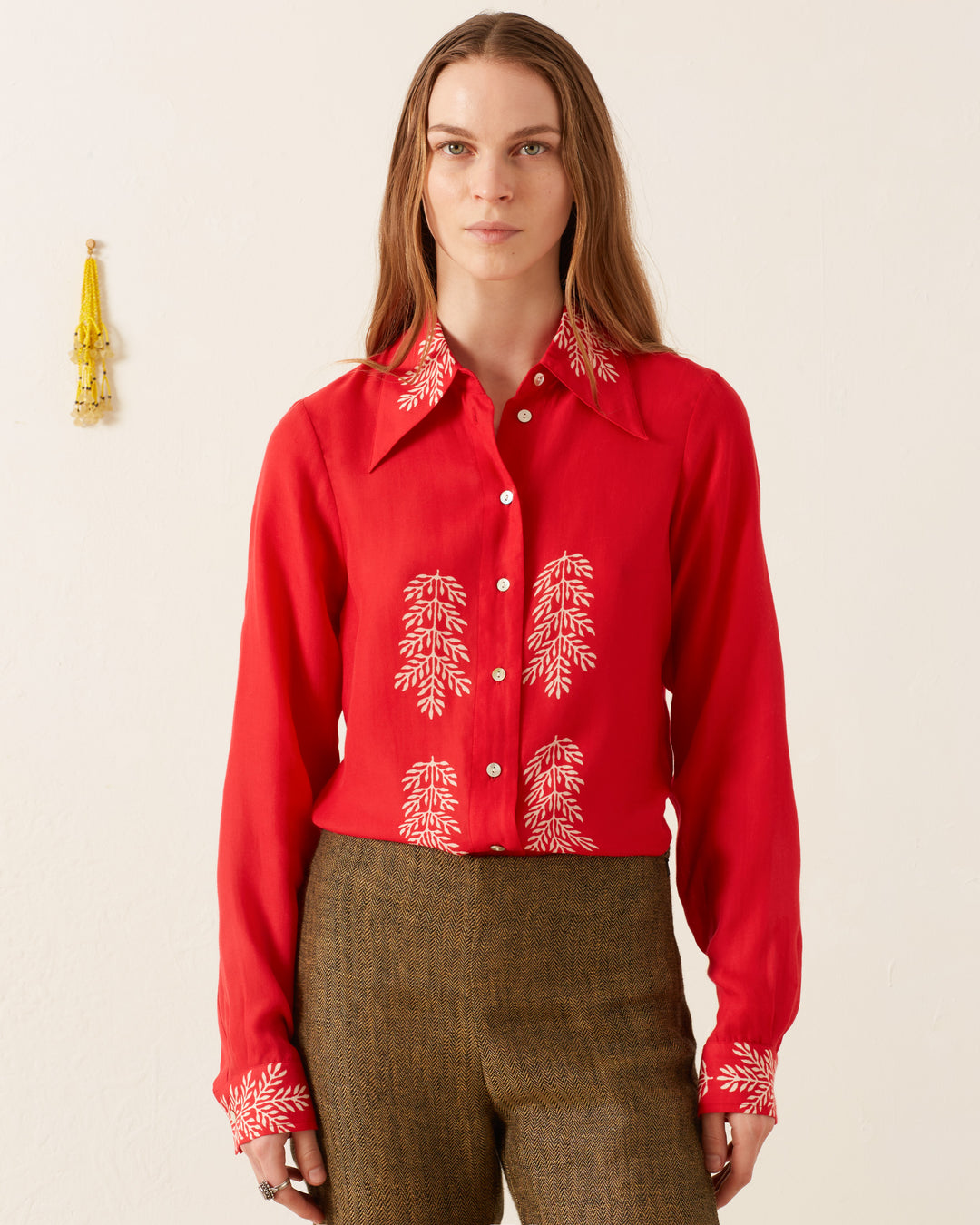 A person with long hair stands against a plain wall, wearing the vintage-inspired Dolly Cherry Mint Shirt, which is adorned with white leaf patterns on the collar, chest, and cuffs. Paired with brown pants, their style is subtly complemented by a keychain hanging in the background.