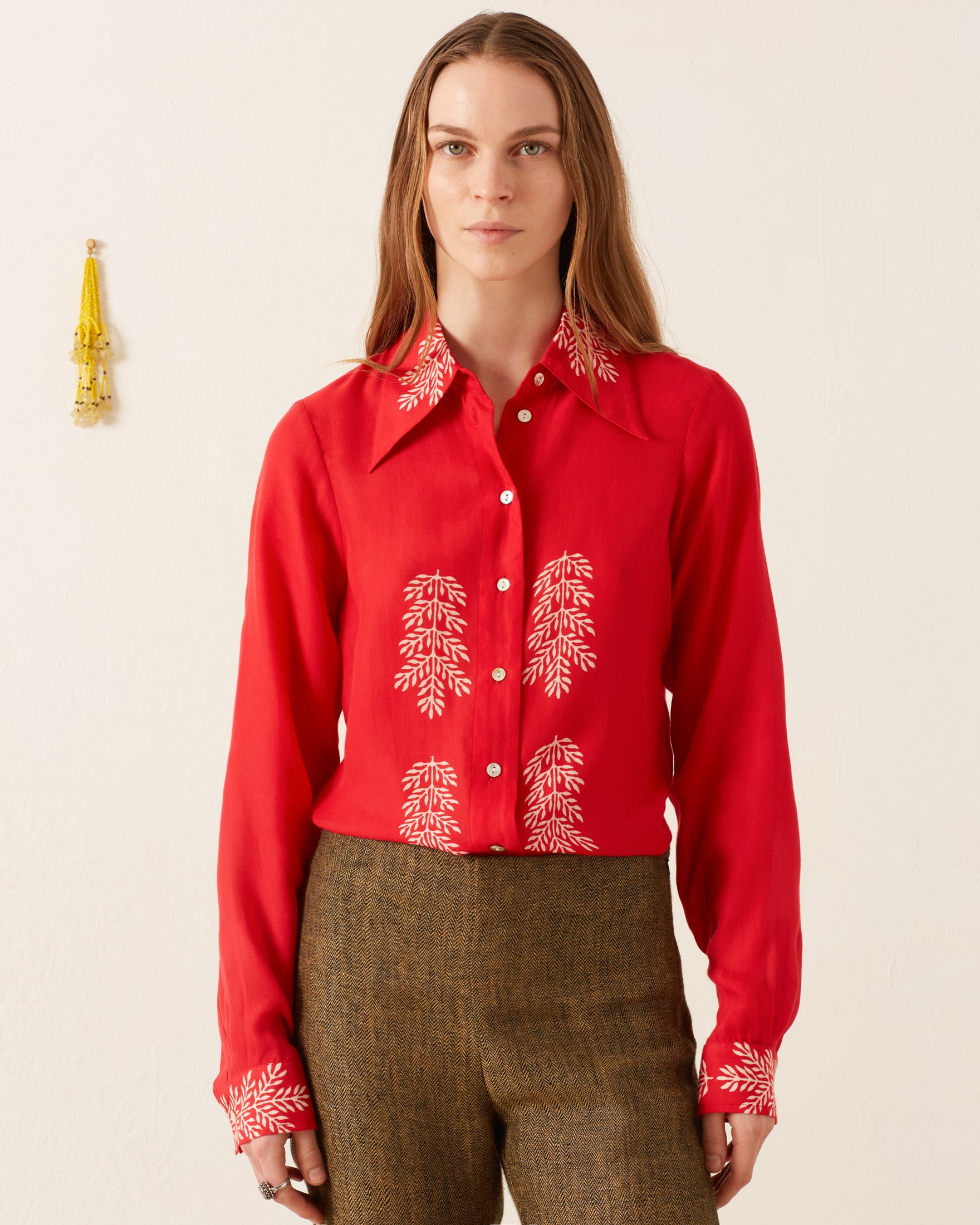 A person with long hair stands against a plain wall, wearing the vintage-inspired Dolly Cherry Mint Shirt, which is adorned with white leaf patterns on the collar, chest, and cuffs. Paired with brown pants, their style is subtly complemented by a keychain hanging in the background.