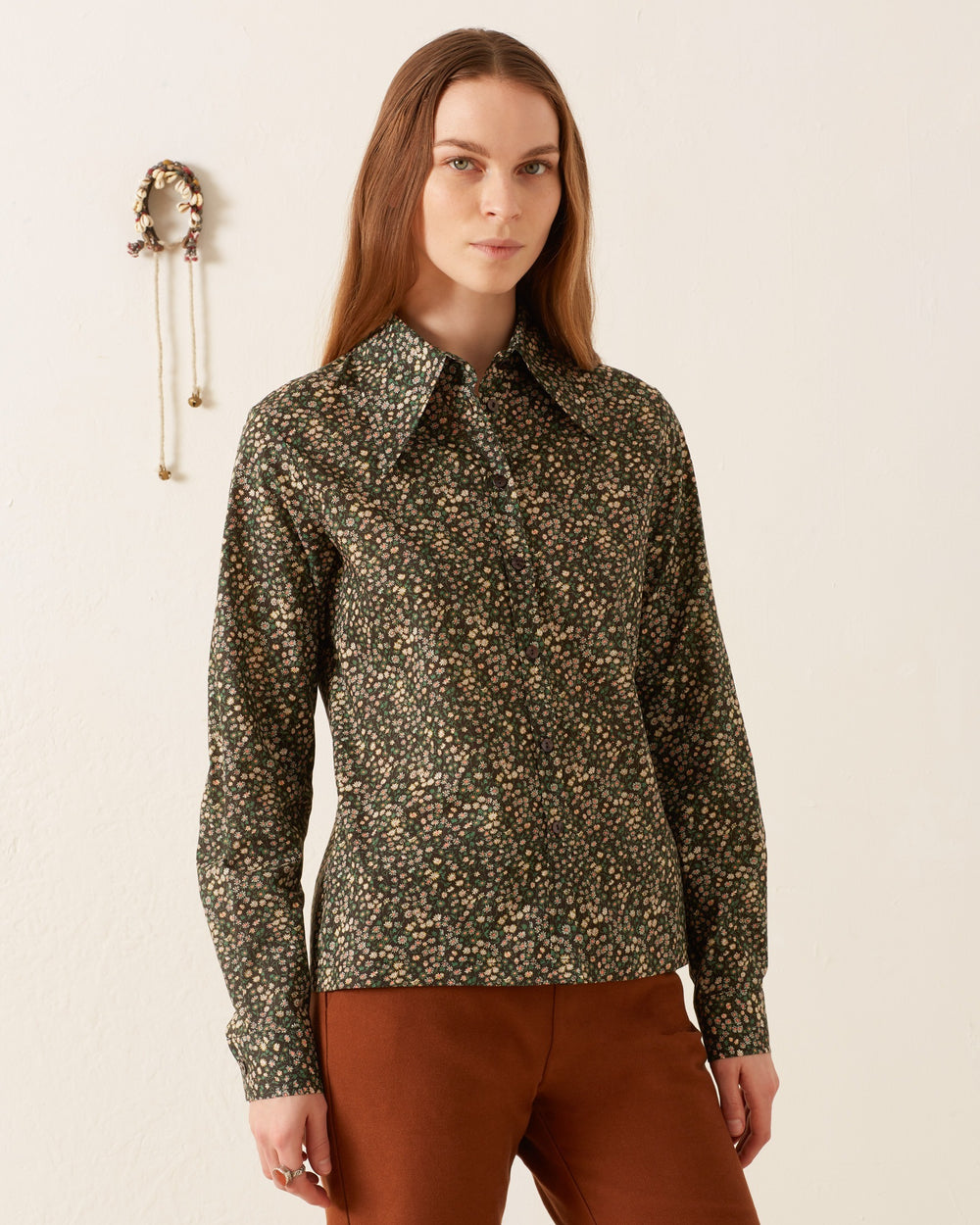 A person with long hair wears a vintage-inspired, dark Dolly Moonflower Shirt and brown pants as they stand in front of a white wall.