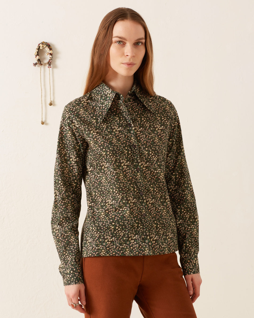 A person with long hair wears a vintage-inspired, dark Dolly Moonflower Shirt and brown pants as they stand in front of a white wall.