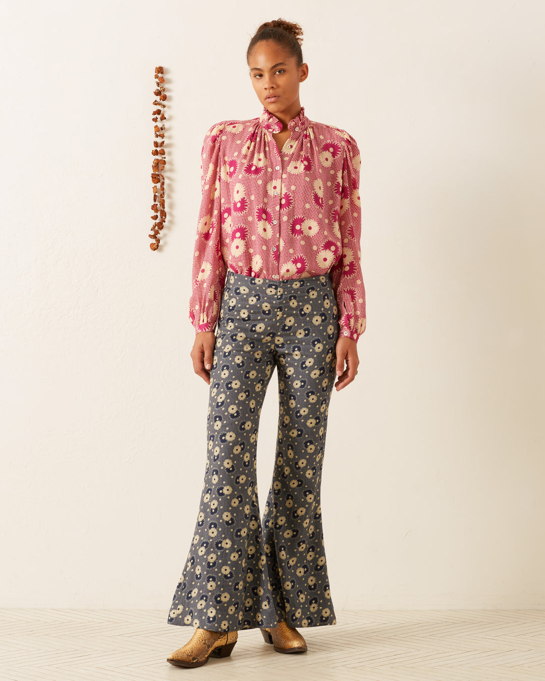 A person stands against a white wall wearing an Annabel Berry Daisy Shirt paired with blue floral bell-bottom pants. They are also sporting brown shoes. A vertical string of small objects hangs on the wall beside them, adding an intriguing touch to this vintage-inspired scene.