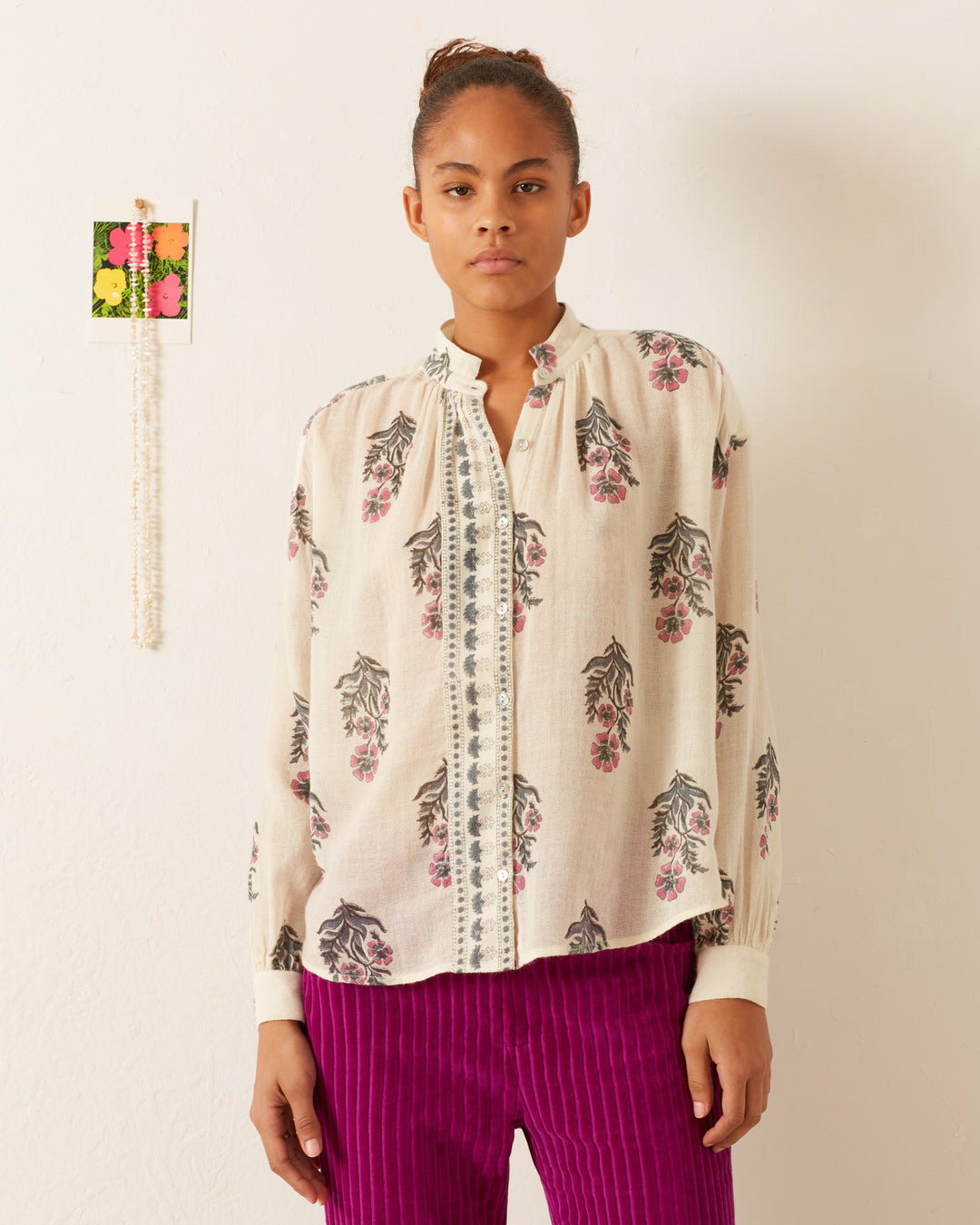 A person stands against a plain wall, wearing the Kiki Mirabilis Wool Shirt, which features a light, vintage-inspired floral motif with decorative front trim, paired with vibrant purple pinstriped pants. A small flower picture and a pink string adorn the wall beside them.