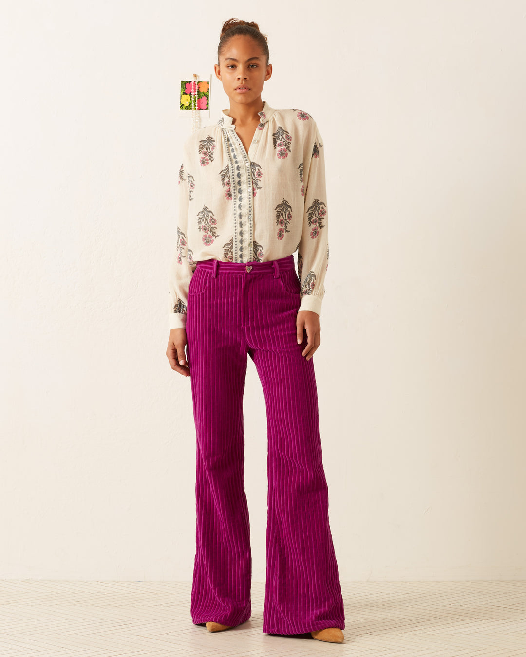 A person stands indoors against a plain white wall, wearing the Kiki Mirabilis Wool Shirt, a vintage-inspired blouse adorned with hand-block printed floral motifs, paired with vibrant magenta flared pants. They are looking forward, with their hair tied up and hands resting by their sides.