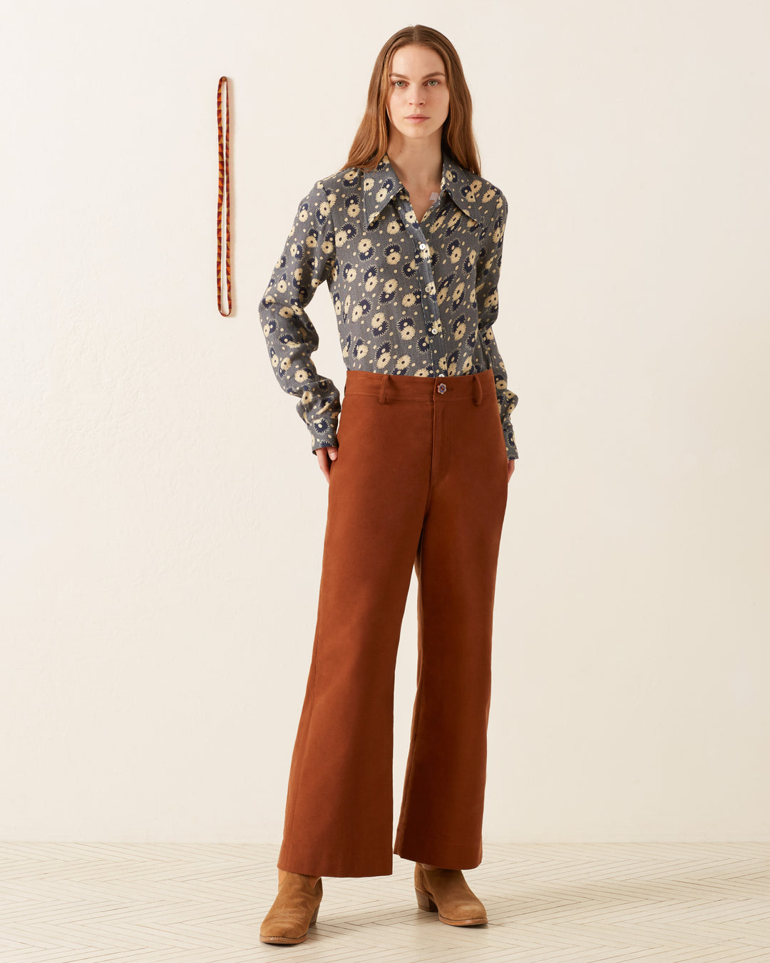 A person stands against a beige wall, wearing a vintage-inspired Dolly Navy Daisy Shirt paired with brown wide-leg pants and tan boots. A rope hangs on the wall beside them.