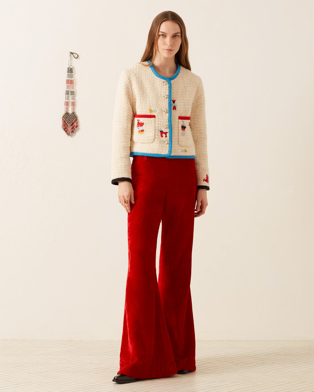 A woman stands against a plain background, wearing a cream jacket with colorful trims and the Charlie Cherry Velvet Pant in '70s-style red velvet. A decorative wall hanging is visible to her side.