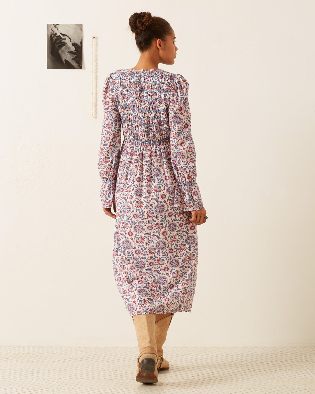 A person stands facing a white wall, wearing the Sabine Amazon Floral Dress, which is a vintage-inspired maxi dress featuring pink and purple floral patterns, long sleeves, and a hand-smocked bodice. They have paired it with tan boots. A small photo is pinned to the wall above the person's shoulder.