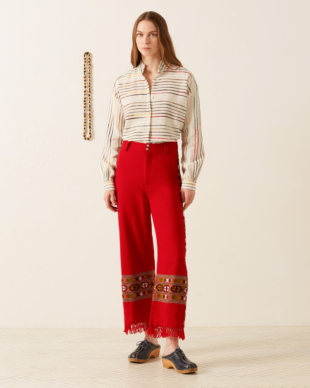 A woman stands against a light background, wearing the Scout Scarlet Blanket Pant, which features intricate Himalayan weaving traditions near the hem and pairs beautifully with a striped blouse. She has long hair and is wearing black shoes, while a decorative wall piece hangs in the background.