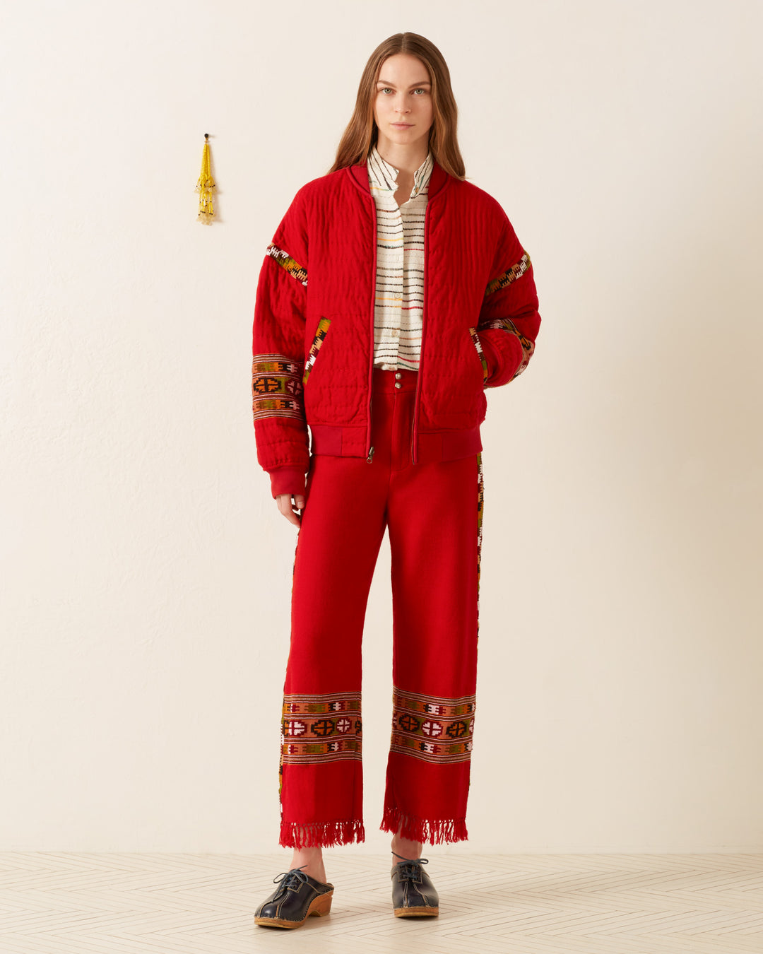 A person stands against a plain backdrop, wearing the Scout Scarlet Blanket Pant, characterized by intricate patterns and influenced by Himalayan weaving traditions. The pants feature fringe at the hem and are paired with a striped shirt and black shoes, with a small decorative object hanging on the wall behind.