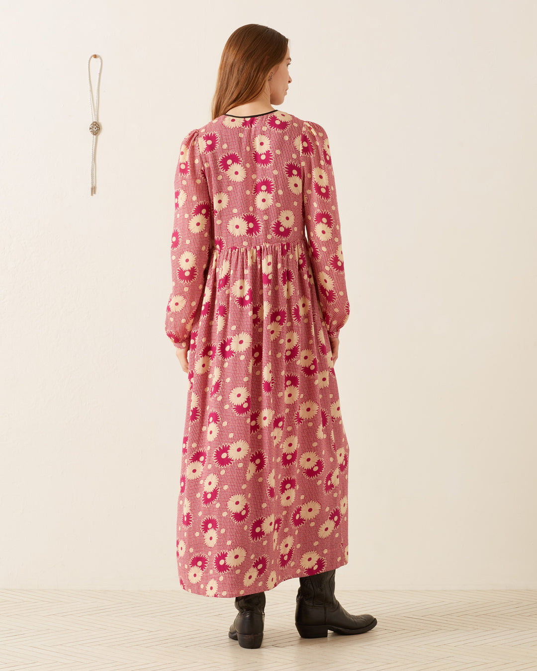 A person with long hair stands facing away in a Winifred Berry Daisy Dress, featuring a V-neckline and retro floral prairie design. The romantic silhouette showcases white and black flowers against pink fabric. They pair the dress with black boots while standing in a light-colored room adorned with simple wall decor.