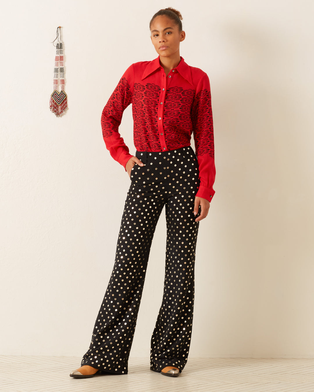 A person stands against a plain white wall, wearing a red long-sleeve shirt with a lace pattern and the Charlie Stardust Pant, which are black wide-leg pants accented with white polka dots. A decorative wall hanging is visible on the left. The person's hair is tied back, enhancing the elegance of the ensemble featuring subtly embellished pants.