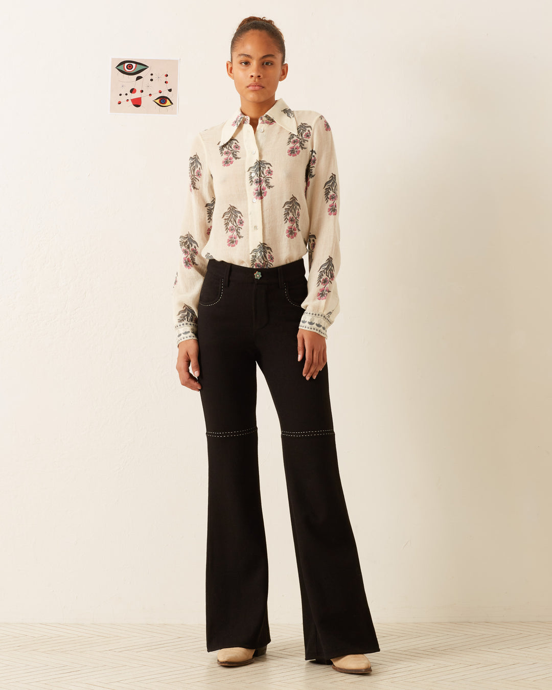 A person stands against a white wall wearing the Dolly Mirabilis Wool Shirt, featuring a vintage-inspired floral motif and long sleeves. The shirt is tucked into high-waisted black flared pants, complemented by beige shoes. Behind them hangs a small, abstract artwork adorned with eyes and patterns.