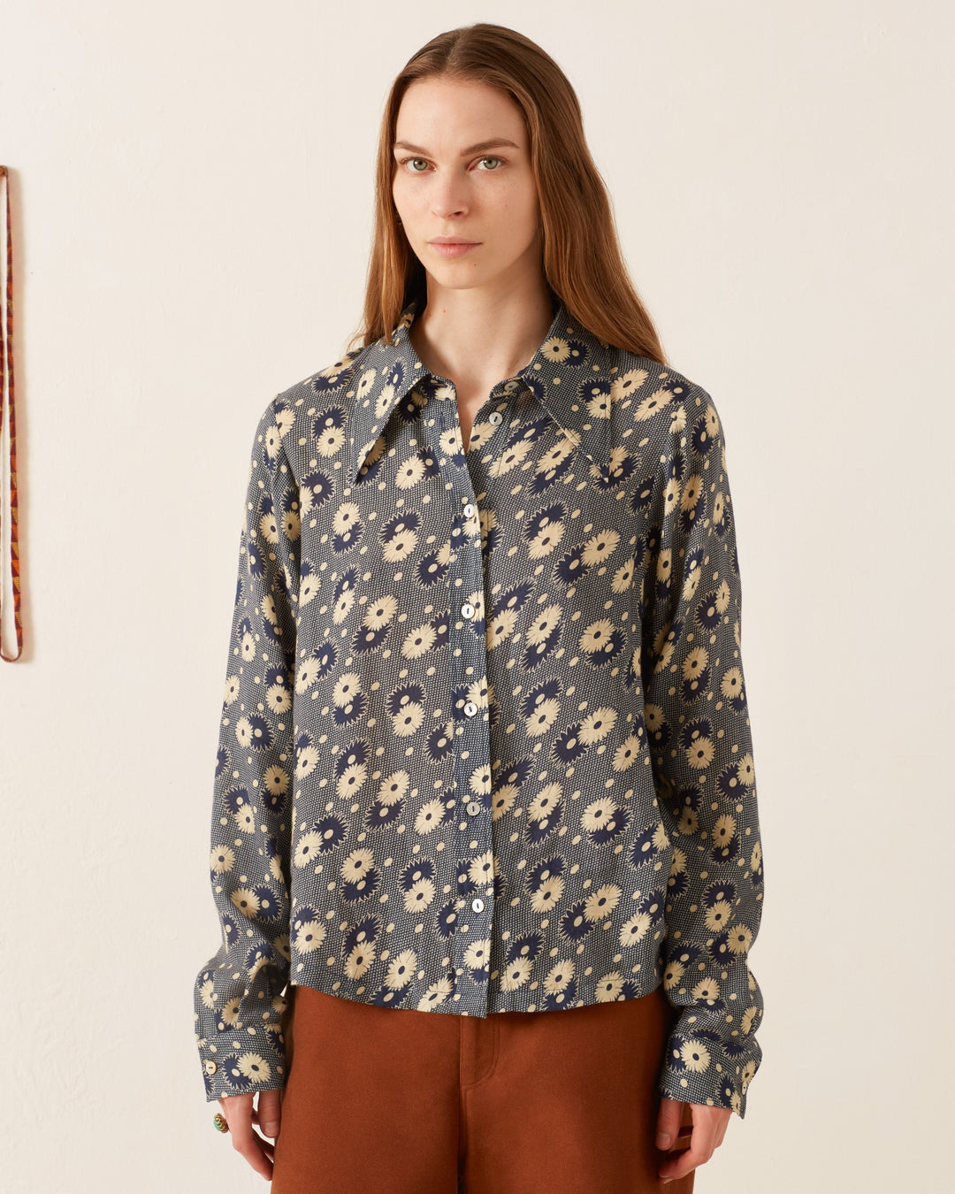 A woman with long brown hair wears a Dolly Navy Daisy Shirt and brown pants, standing in front of a plain background.