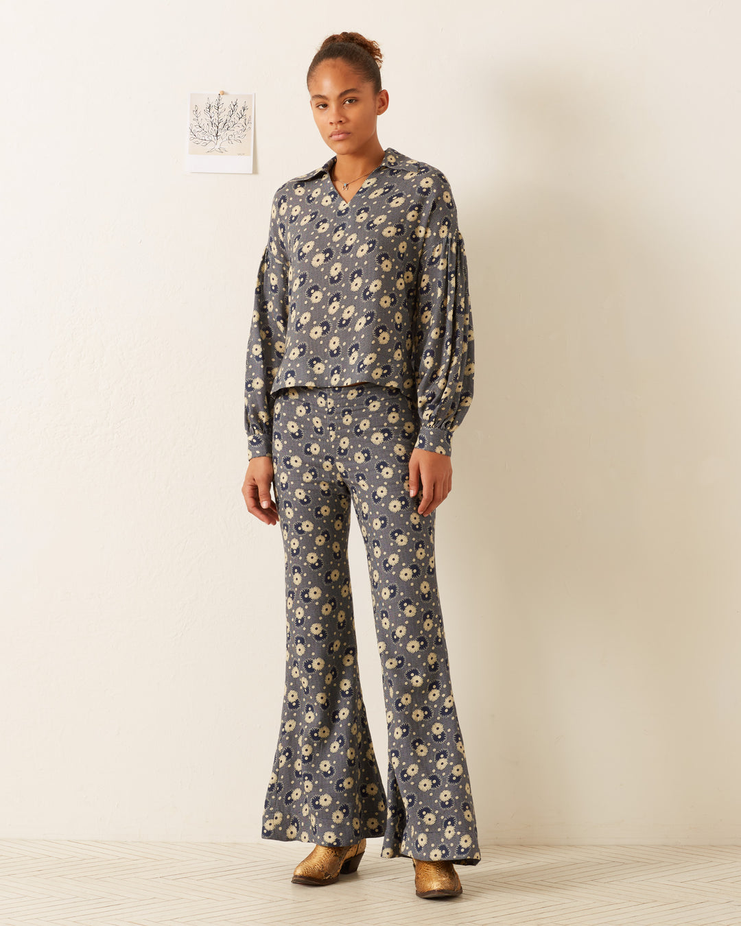 A person stands in the room, showcasing a '70s-style floral print ensemble featuring the Farah Navy Daisy Pant with bell-bottom flare. Brown shoes complete the look, while in the background, a small piece of art adds to the retro charm.