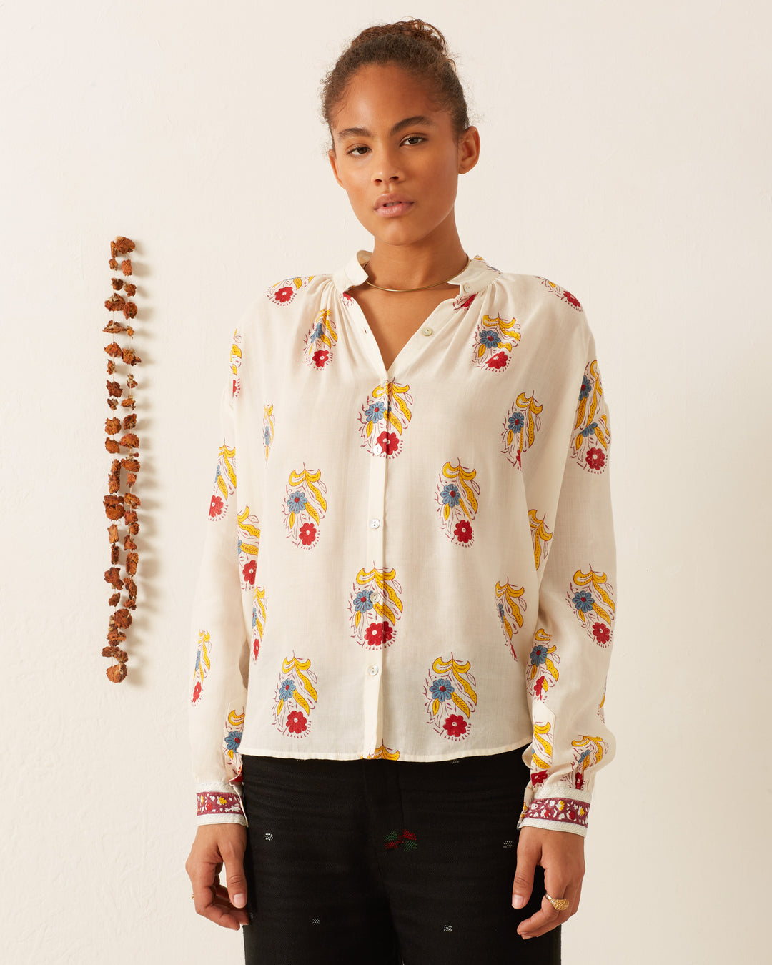 A person wearing the Kiki Fireflower Shirt, adorned with colorful floral motifs and paisley patterns that are hand-block printed on Tencel twill, stands against a plain wall. They have a calm expression, and a string of small, dried objects hangs beside them.