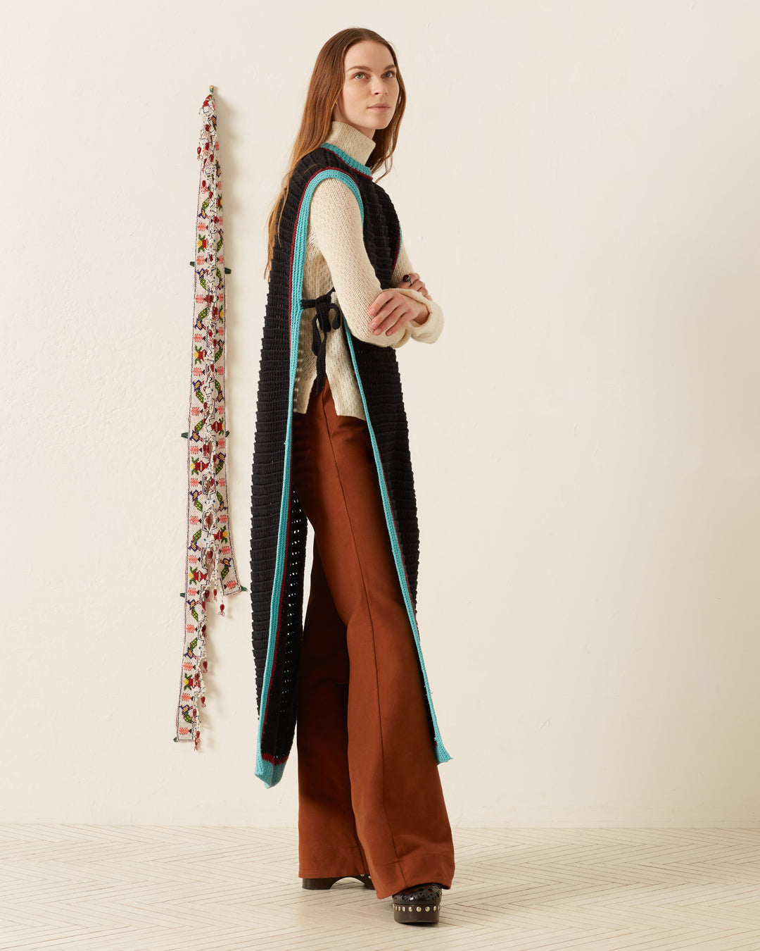A person stands sideways in a minimalist room wearing the Omer Equinox Crochet Top— a vintage-inspired black knit vest with blue trim— over a cream turtleneck sweater and brown wide-leg pants.