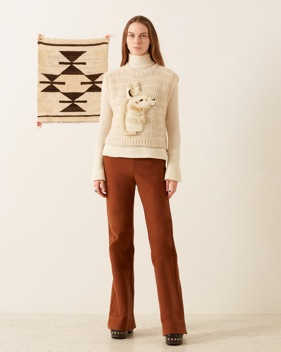 In a cozy room featuring an indigenous geometric patterned textile on the wall, a woman dons the Oona Llama Crochet Top—a vintage-inspired cream knitted vest showcasing a llama design. She pairs it with a beige long-sleeve top and rust-colored pants, all complemented by the warm wooden flooring.