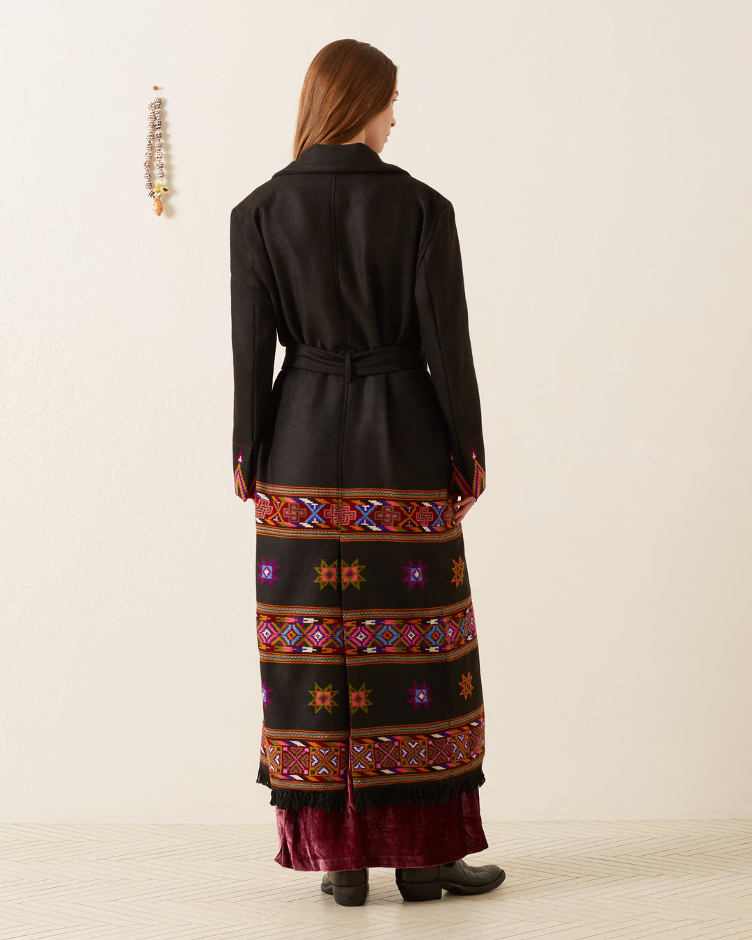 A woman stands with her back to the camera, draped in the Penelope Wildflower Wool Coat, a long black wool coat adorned with colorful embroidered patterns at the hem. Her brown hair cascades gently as she poses against a neutral background, highlighted by a beaded decoration hanging on the wall.