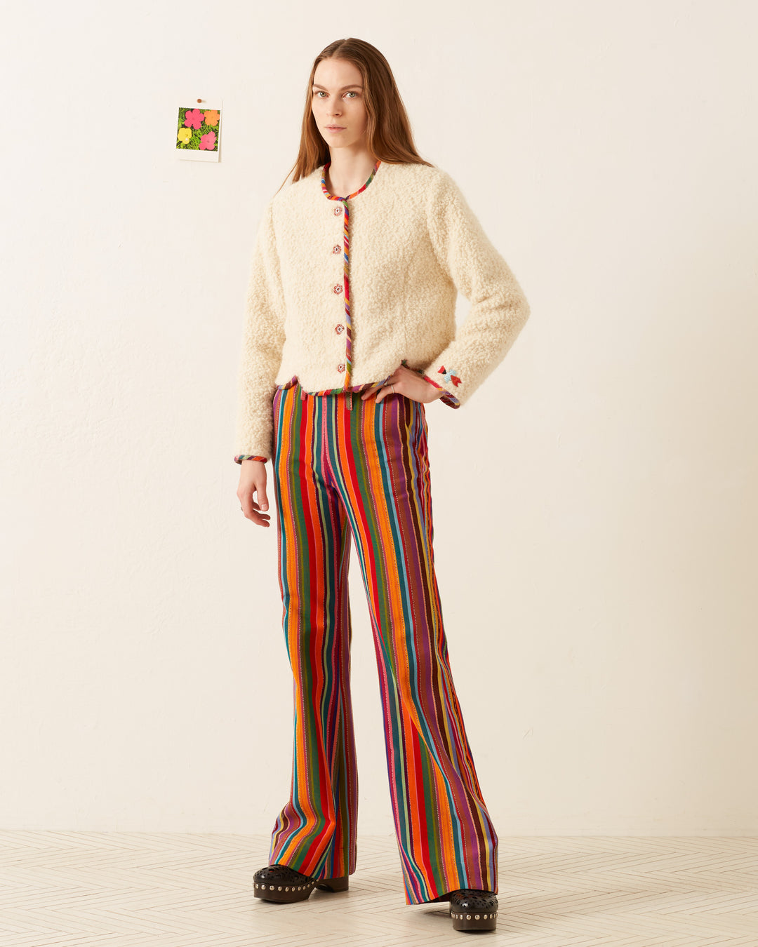 A person stands against a light background wearing the Rowan Suri Wool Jacket adorned with floral buttons and vibrant, striped flared pants in red, orange, and green. Black platform shoes complete the outfit. A small, colorful artwork is on the wall behind them.