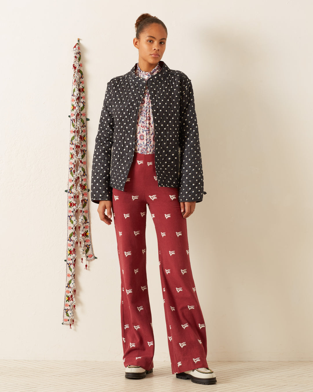 A person wearing a Tamil Raven Dot Silk Jacket, a patterned shirt, and red patterned pants stands against a plain wall. A colorful hanging decoration is on the left.