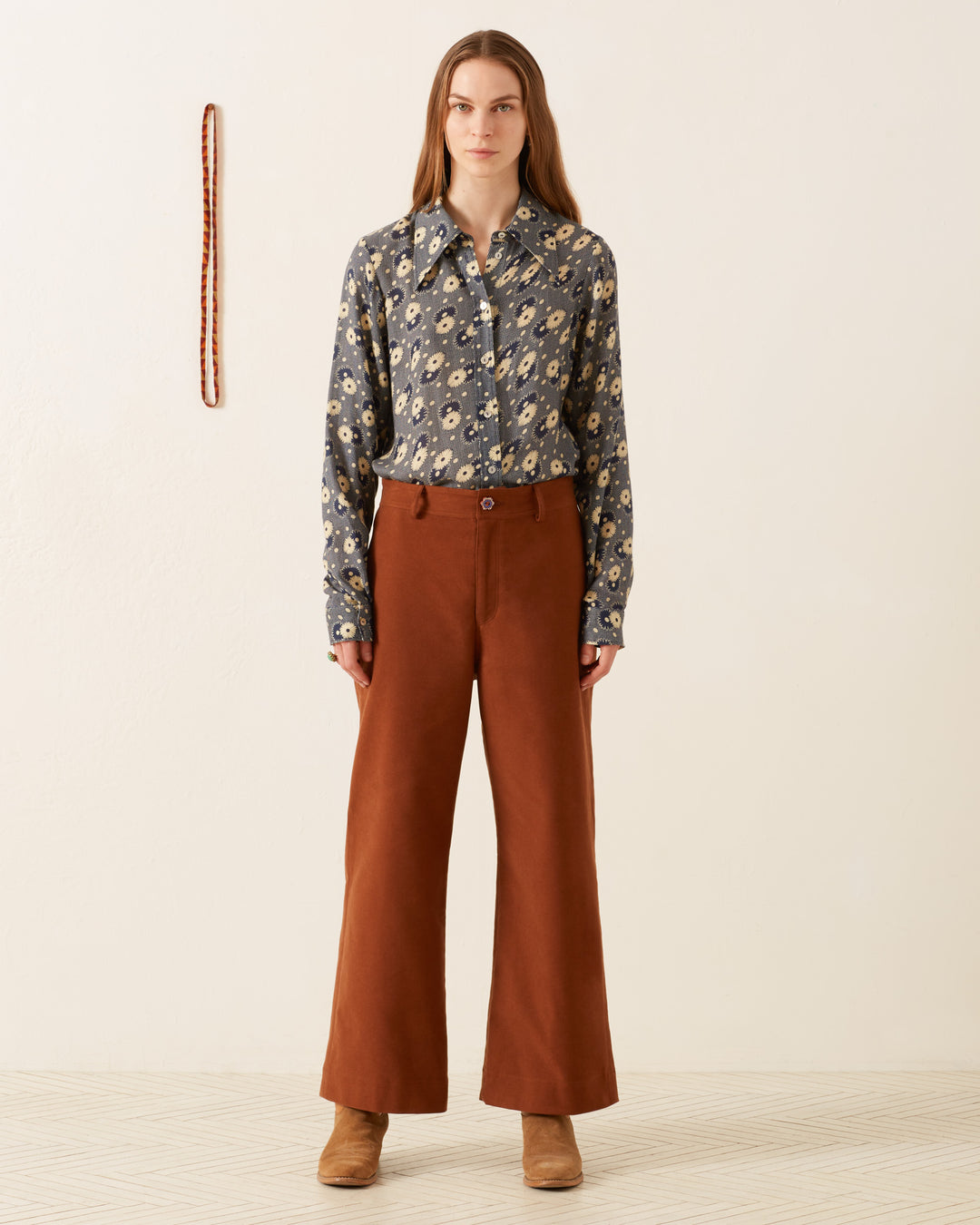 A person stands against a light background wearing a Dolly Navy Daisy Shirt, brown pants, and boots. A thin, vertical object is mounted on the wall in the background.