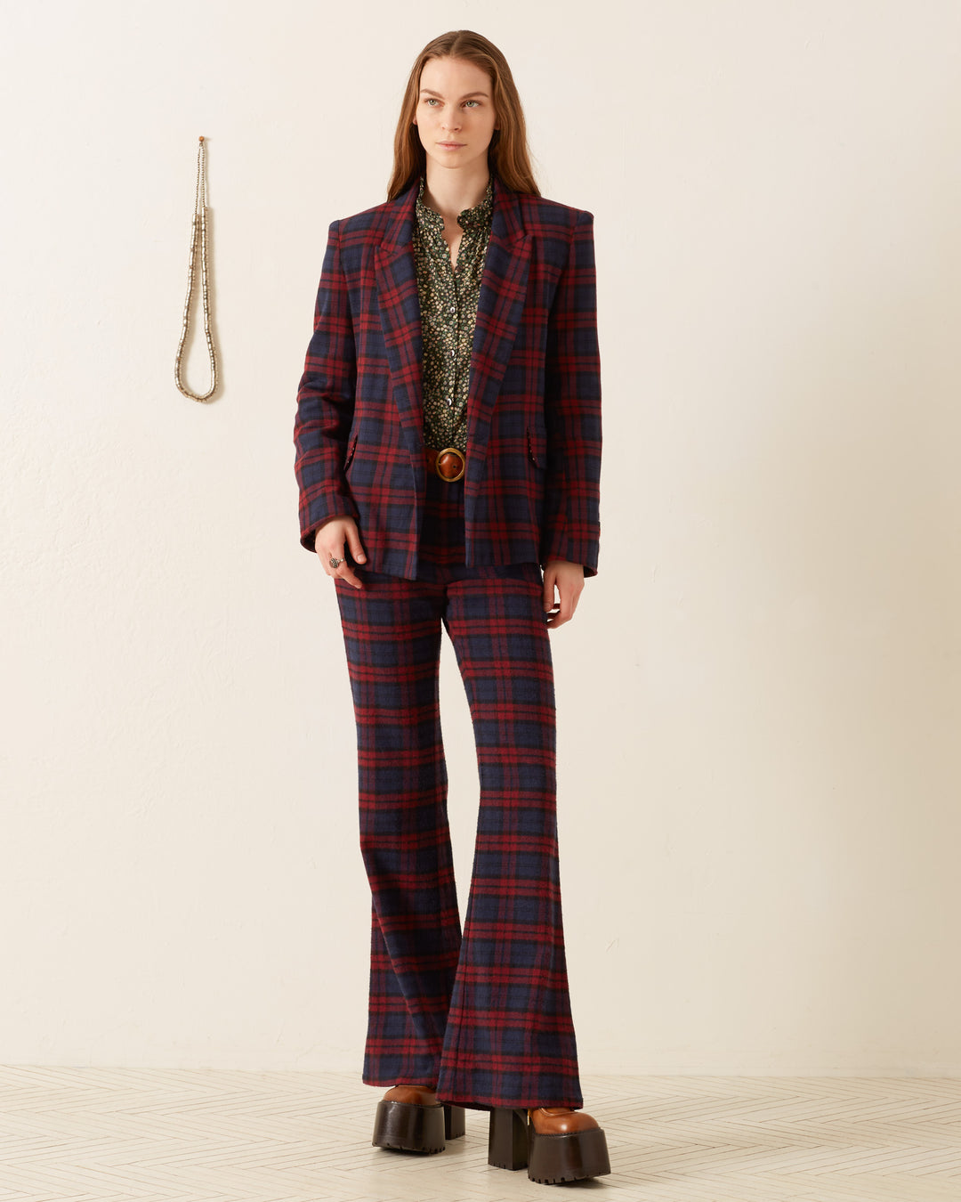 A person stands against a light background wearing the Dree Berry Plaid Blazer, a patterned shirt, and platform shoes. A necklace is hanging on the wall.