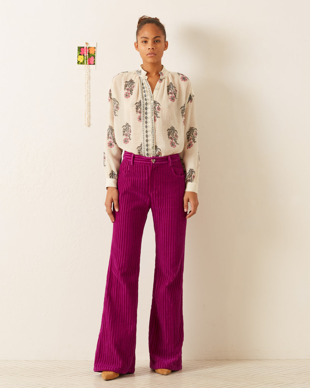 A person stands against a white wall wearing the Kiki Mirabilis Wool Shirt, which features a vintage-inspired floral motif, paired with vibrant magenta flared pants. A small decorative object and a string of beads hang on the wall beside them.