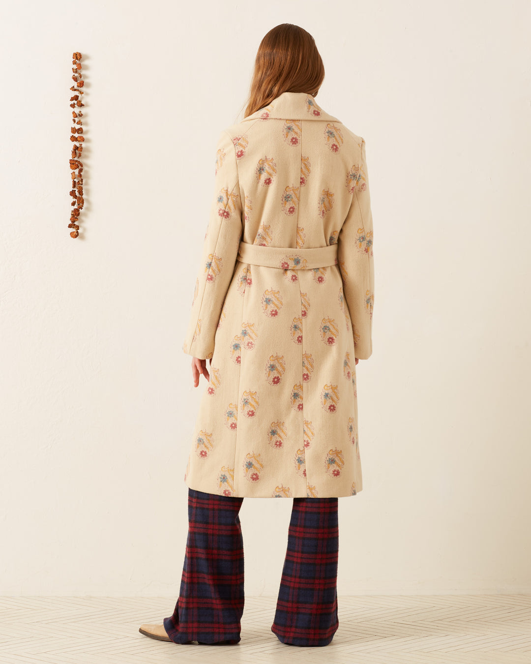 Standing in front of a cream-colored wall, a person with long hair wears the Officer Fireflower Wool Coat, which features a floral pattern, along with plaid pants. The vertical line of small brown decorations enhances the hand-block printed designs on the coat, while the floor displays a subtle geometric pattern.