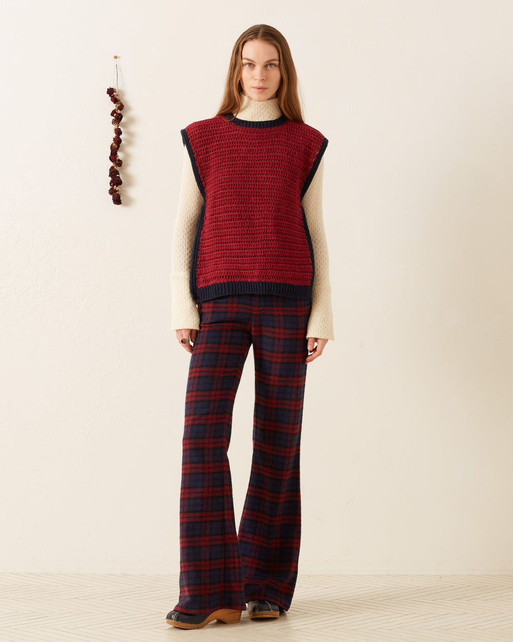 A woman stands indoors wearing the Oona Mulberry Crochet Top over a cream long-sleeve, paired with red and navy plaid flared pants. Dried flowers are displayed on the wall behind her.