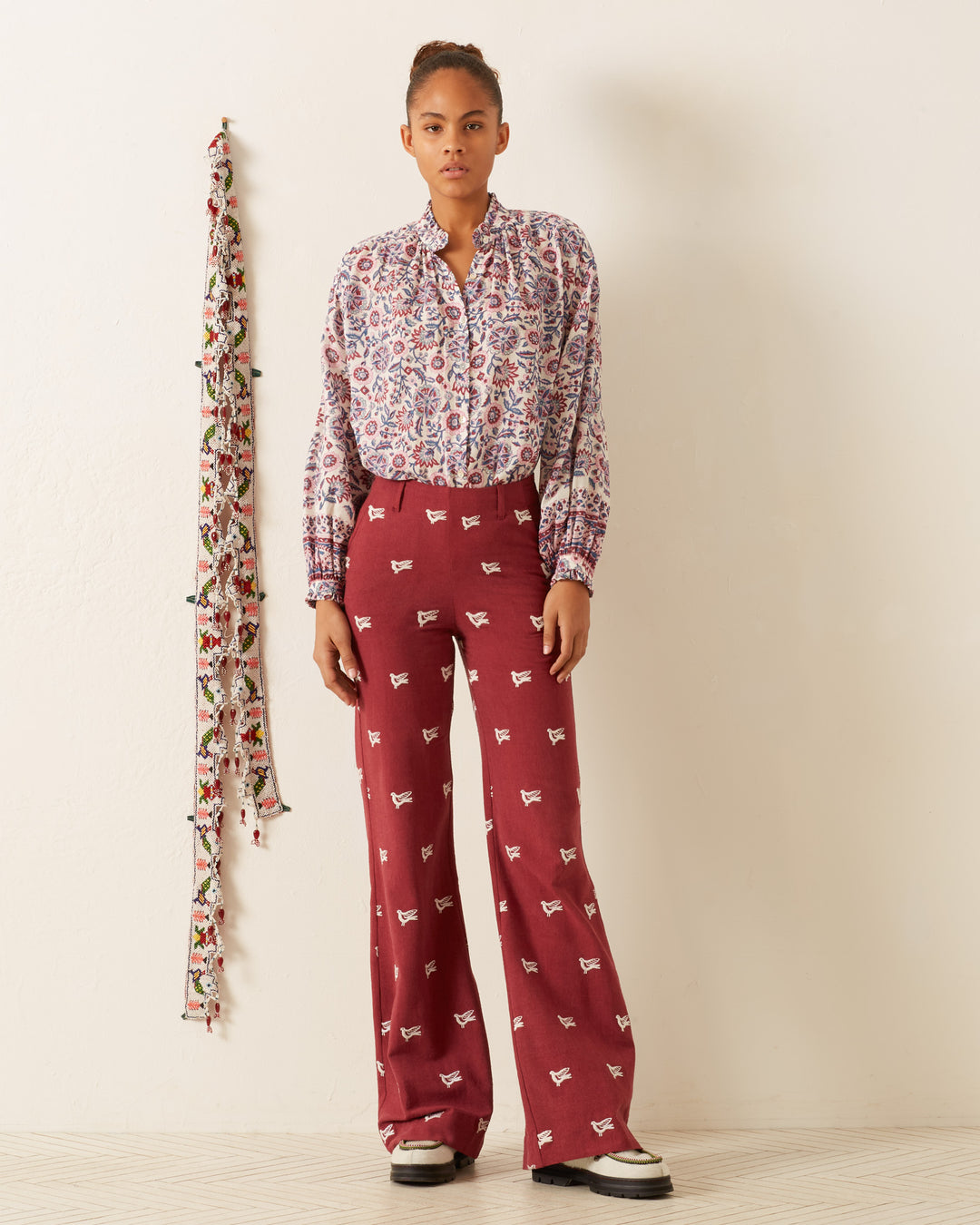 Against a light wall, a person stands wearing the Poet Amazon Floral Blouse paired with red flared pants adorned with white designs. To their side, a colorful knotted decoration adds charm to the scene. The light-colored floor features subtle stripes, enhancing the overall retro vibe.