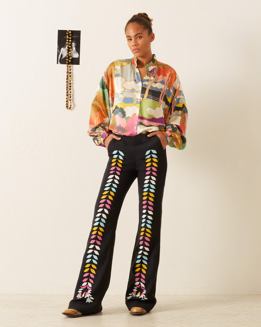 A person wearing a vibrant Poet Aurora Silk Blouse and black pants with multicolored leaf designs stands indoors near a white wall adorned with a small, framed picture.