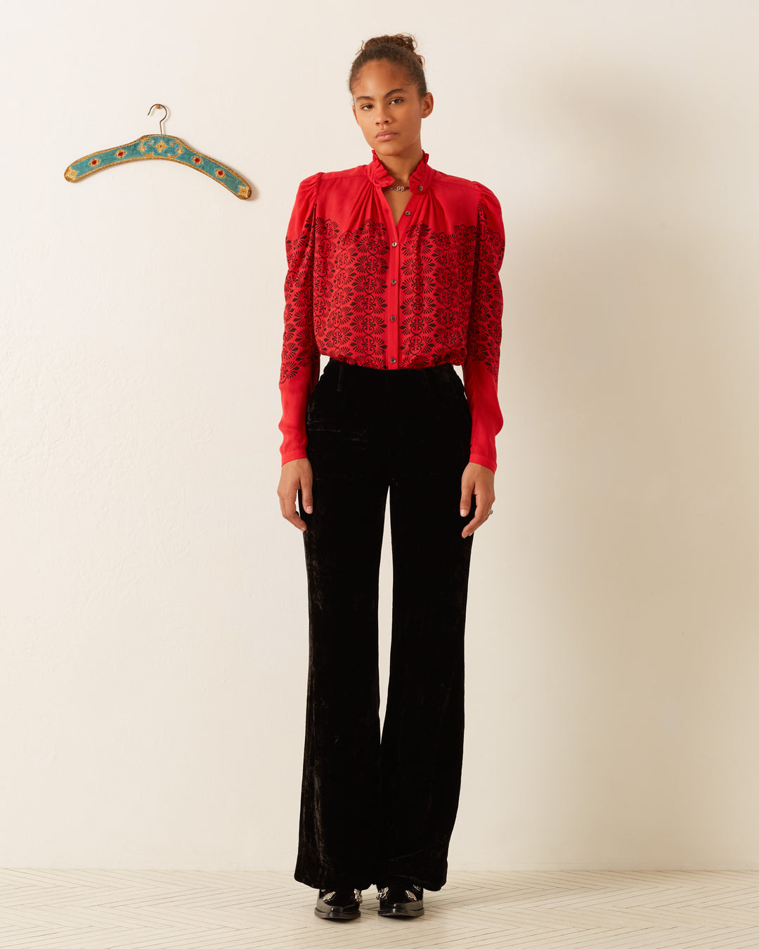 A person stands against a plain background, wearing the Artemis Cherry Trellis Shirt paired with black velvet pants. A decorative hanger is mounted on the wall to the side.