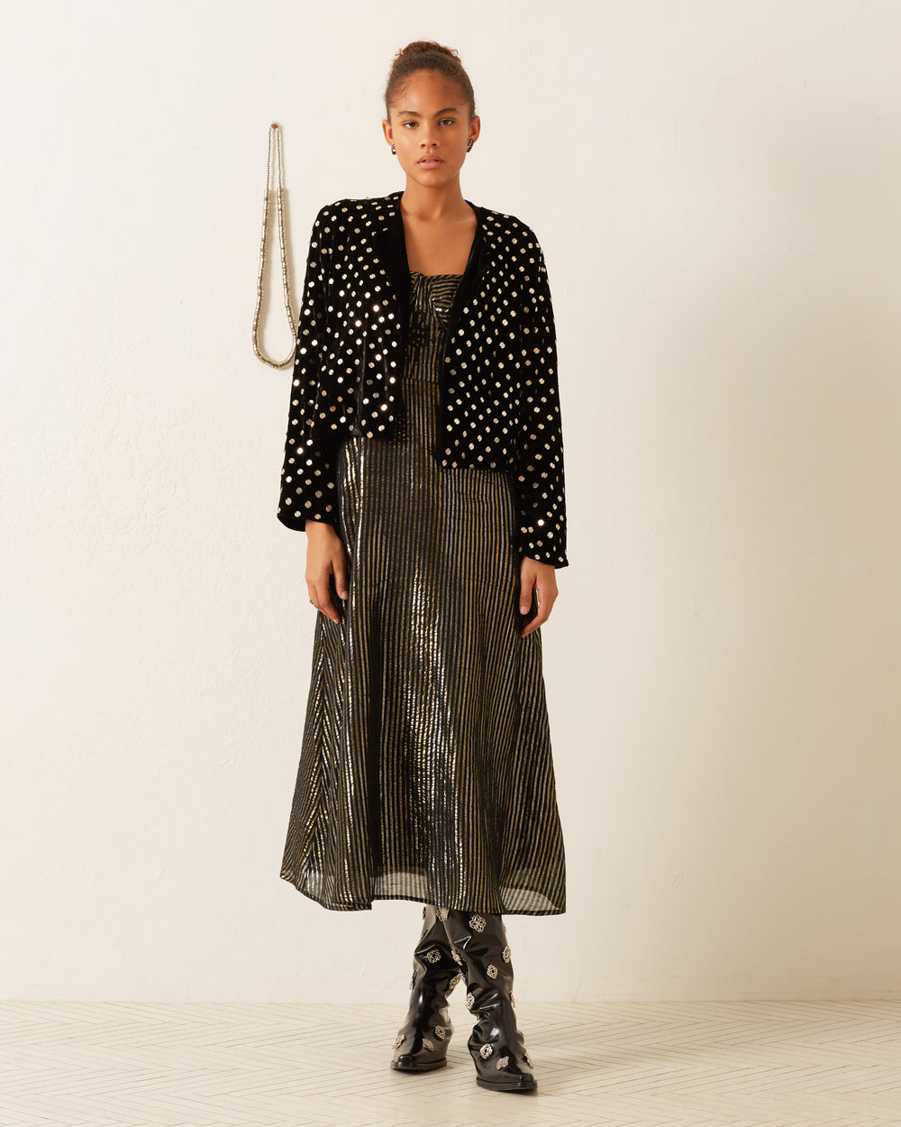 A person wearing the Rowan Stardust Velvet Jacket, featuring hand-embellished black and gold polka dots, a metallic striped dress, and black boots stands in a room with a textured wall and a beaded necklace hanging behind them.