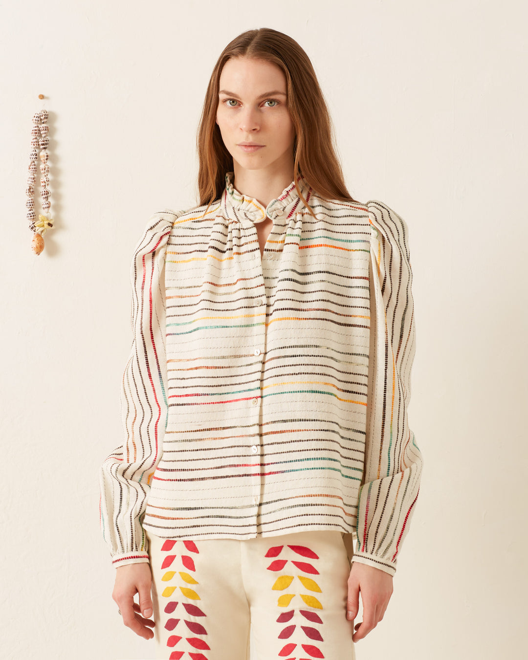 An individual dressed in an Annabel Chroma Weave Shirt and pants featuring leaf designs poses against a simple wall adorned with a string of seashells and beads.