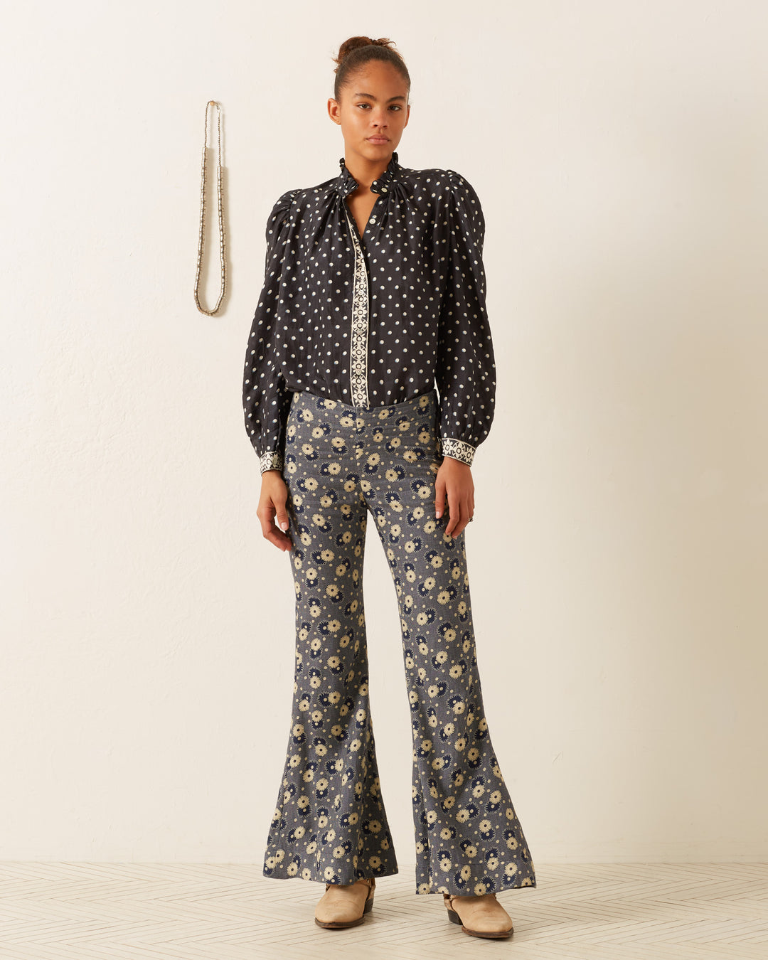 Dressed in the Annabel Raven Dot Silk Shirt and hand-block printed floral flared pants, a woman stands against a white background. A necklace hangs on the wall beside her. With her hair tied up and wearing tan shoes, she embodies a chic blend of patterns with effortless grace.