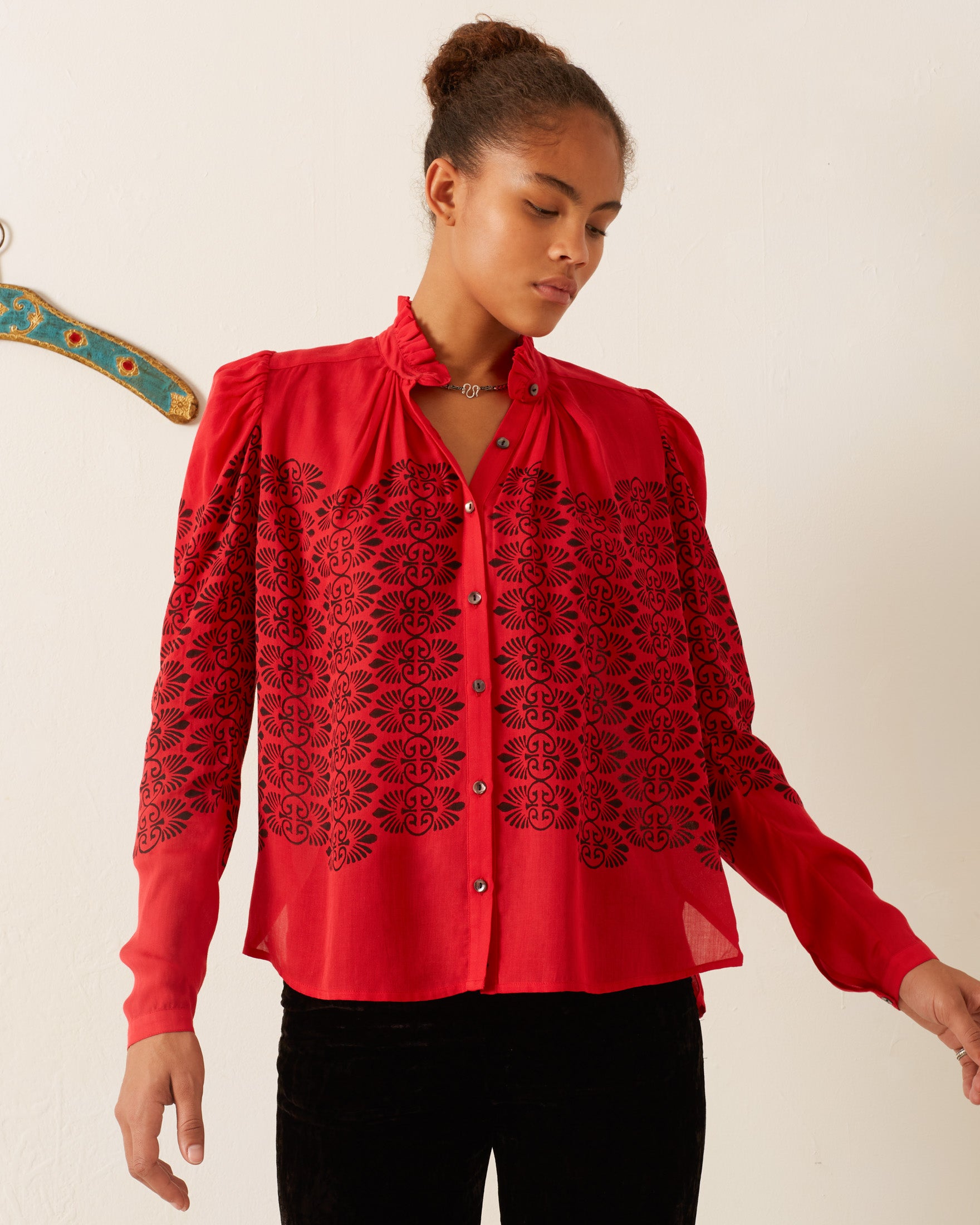 A person wearing the Artemis Cherry Trellis Shirt, characterized by its vintage-inspired design and black floral patterns on a red background, stands against a white backdrop. Made from soft Tencel fabric, the shirt boasts a high collar and button-down front. A decorative wooden hanger is partially visible on the left as the person looks down to their right.