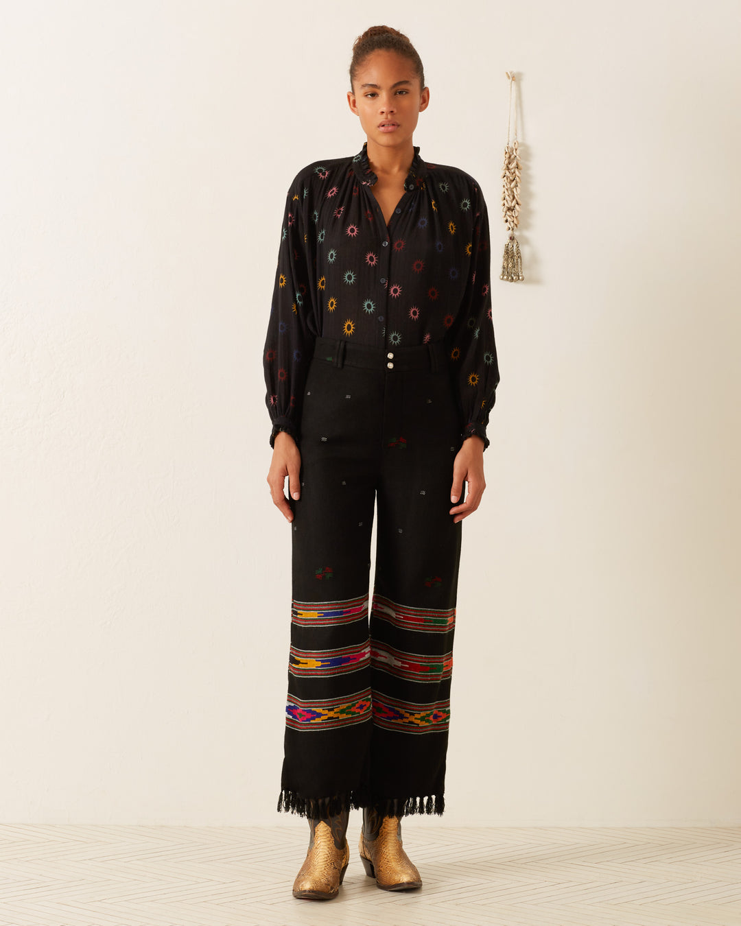 A person wearing the stylish Poet Evening Star Blouse stands elegantly against a white wall. They complement their look with pants featuring vibrant, geometric patterns and brown boots. The decor on the wall subtly enhances the lively outfit in the background.