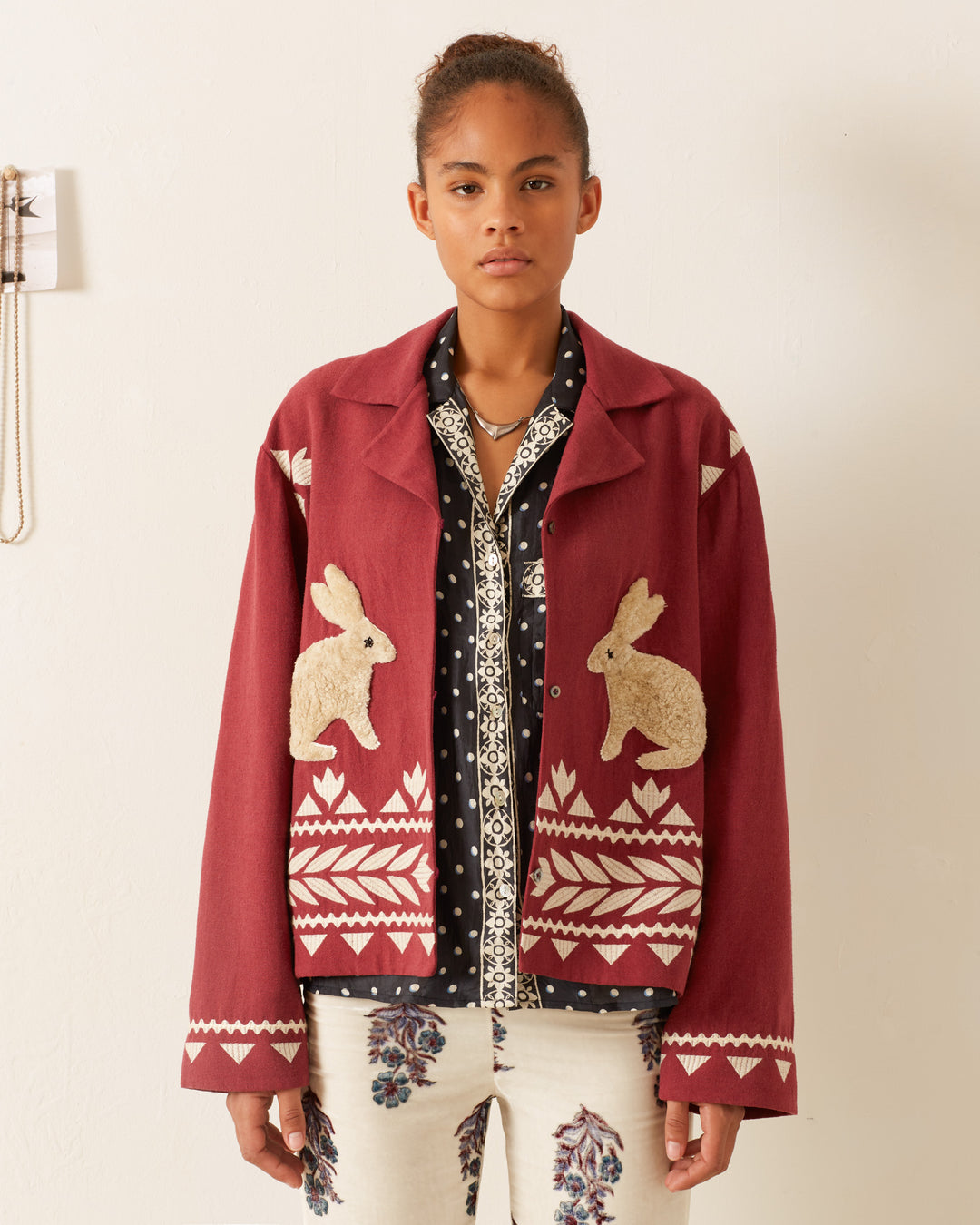 A person wearing the Western Plum Hare Jacket in textured silk, featuring white rabbit designs and geometric patterns, stands against a plain white background. Their curly hair is tied back, and they are sporting a black and white patterned shirt underneath. Their pants display floral patterns.