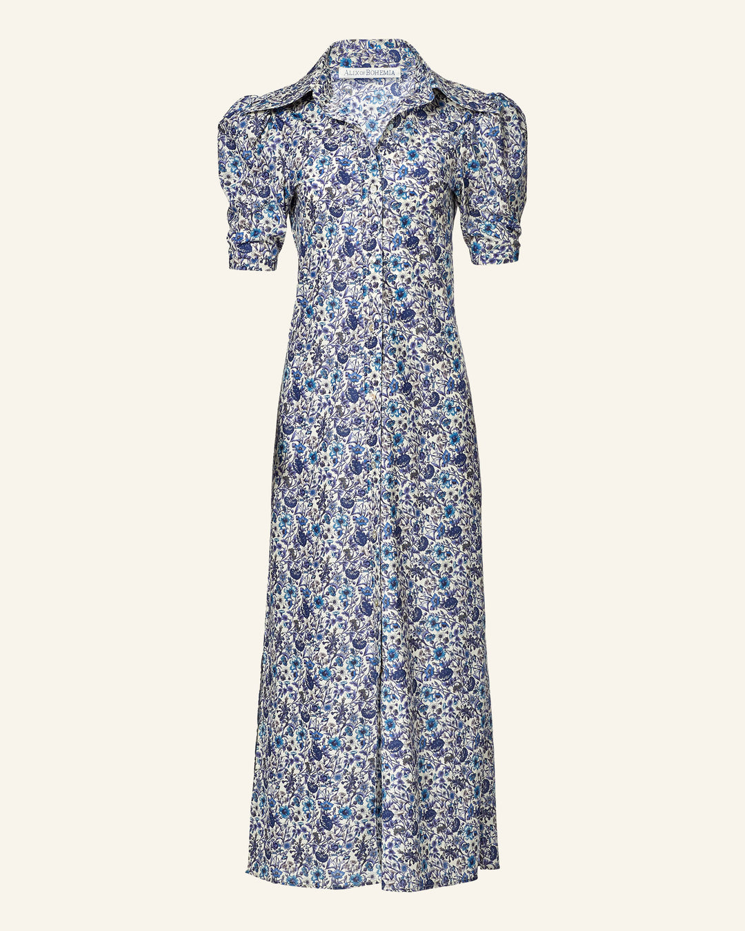 The Deneuve Luna Flower Dress is a long Liberty-print dress in blue and white floral on Italian cotton lawn, featuring short puffed sleeves and a collar against a plain, light background.