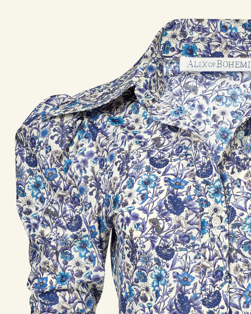 Close-up of the Deneuve Luna Flower Dress showcasing a floral pattern with blue and purple flowers on a white background. The collar is visible and slightly open, revealing a tag reading "Alix of Bohemia.