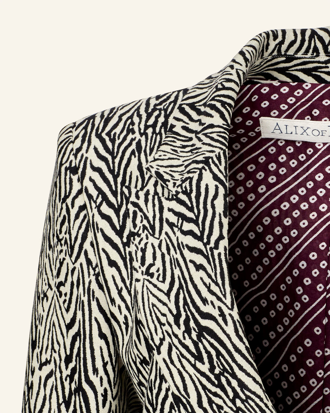 The Dree Zebra Blazer showcases a striking black and white zebra stripe pattern. Its peak lapel adds elegance, and the deep red and white geometric inner lining enhances its style. Crafted from virgin wool, it includes an "ALIXof" label inside.