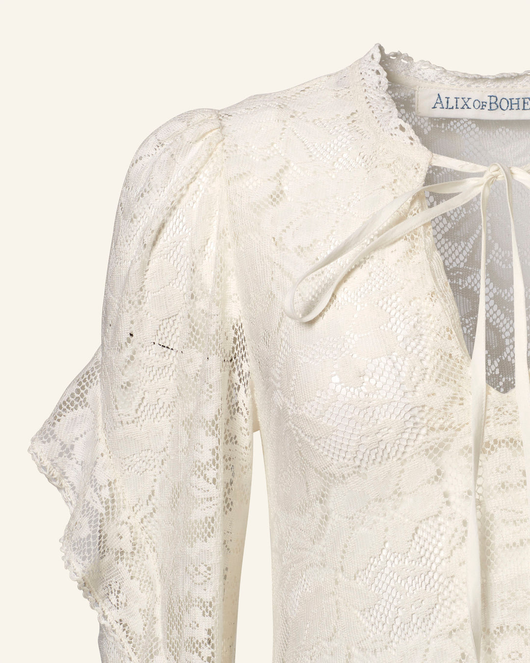 Close-up of a white lace blouse with ruffled sleeves and a tie closure at the neckline, similar to the floral pattern on the Lina Magnolia Lace Dress. The fabric features intricate floral lace. Part of a label reading "ALIX OF BO" is visible, evoking vintage bridal charm.