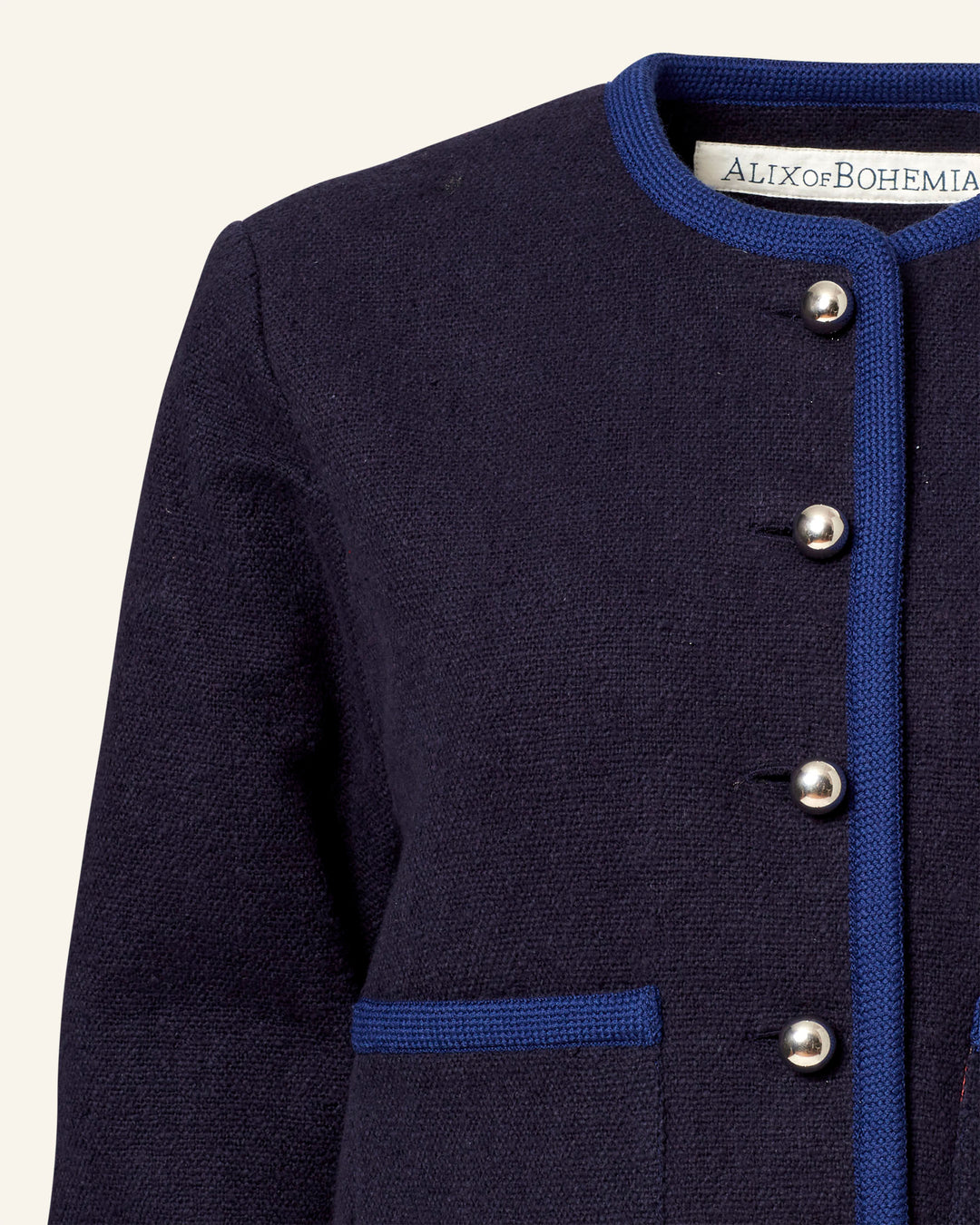 A close-up of the Rowan Midnight Swallow Jacket highlights its textured wool in dark navy with a round neck and blue trim. A row of silver buttons is partially open, revealing a white label that reads "ALIX OF BOHEMIA.