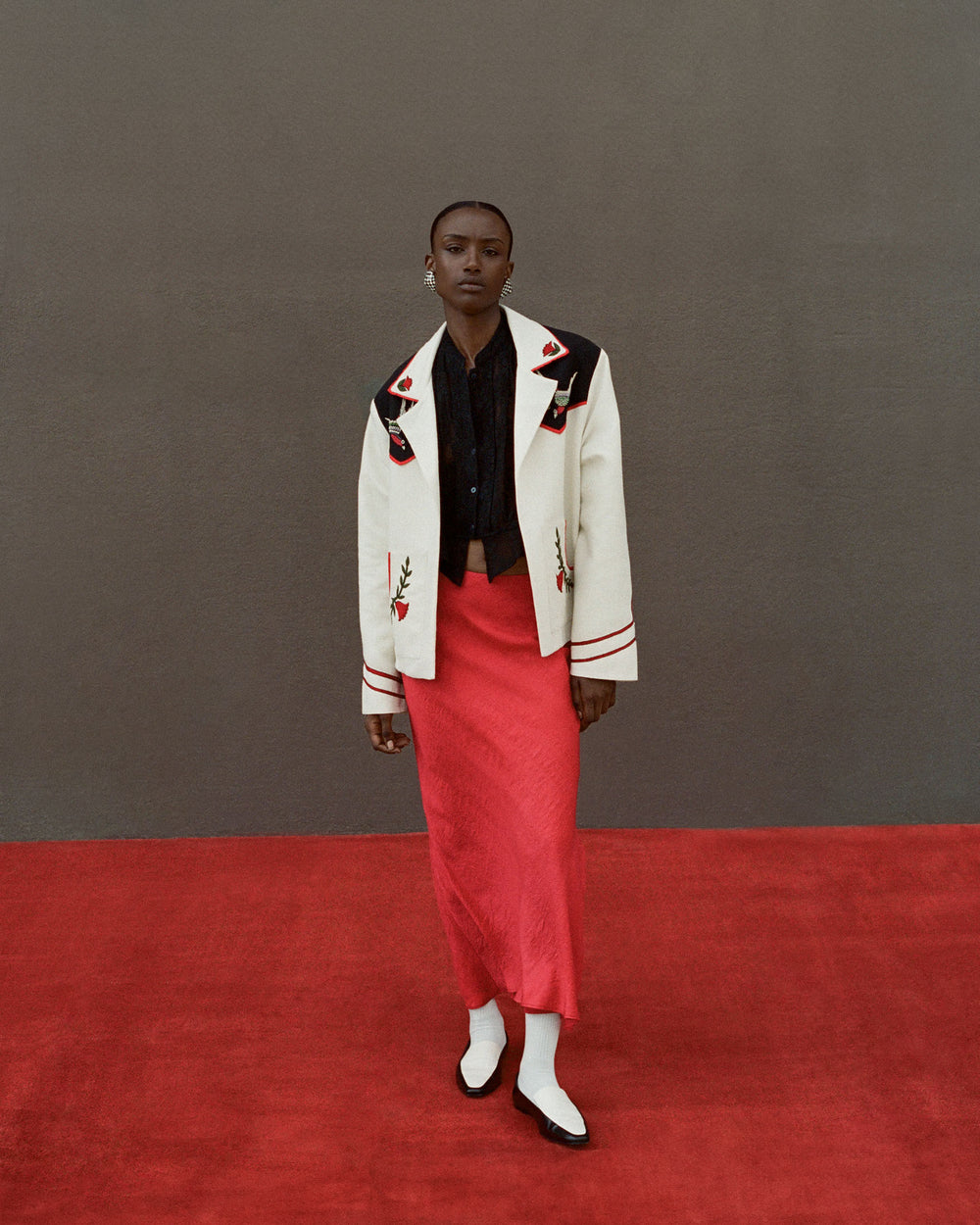 A person stands on a red carpet against a gray background, wearing the Western Royal Flush Jacket, which is silk with floral embroidery. They are also dressed in a black shirt, a red skirt, white socks, and black-and-white shoes.