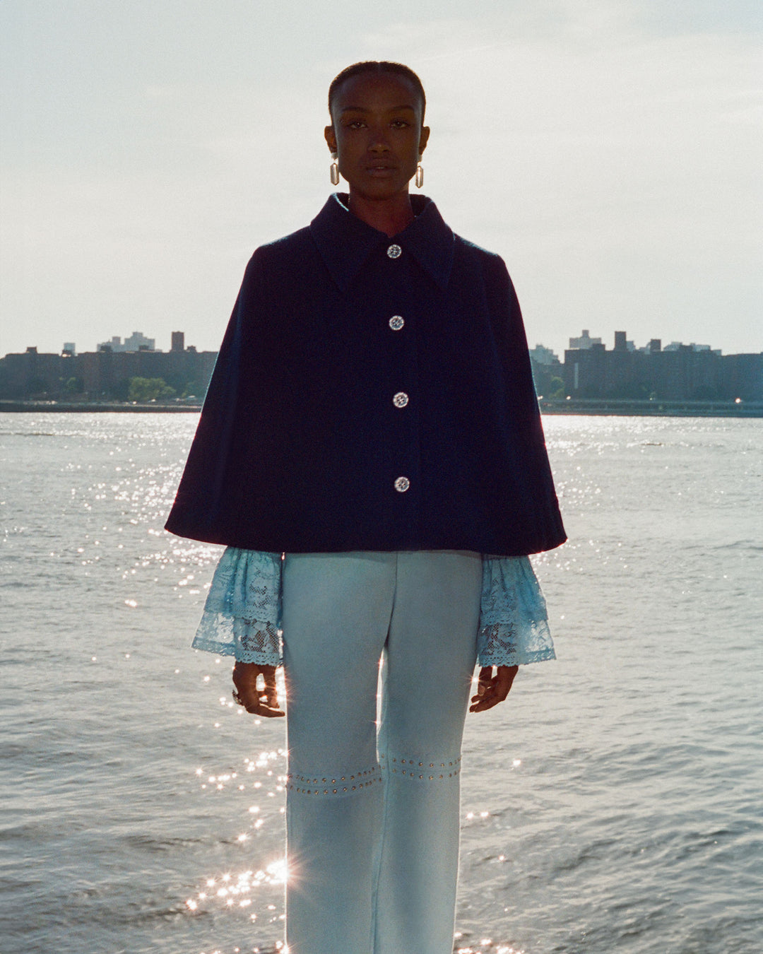 A person stands by a large body of water, draped in a Kensington Midnight Wool Cape with a pointed collar and light-colored pants, as the cityscape stretches in the background.