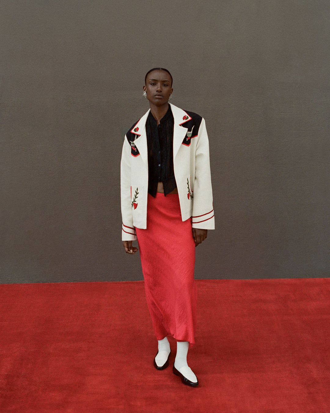 A person stands on a red carpet wearing a Ryland Blackjack Daisy Shirt, a red skirt, and a white jacket with black and red accents. They are also wearing white socks and black loafers, set against a gray wall.