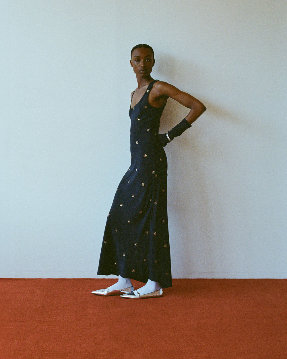 Against a white wall on the red carpet, someone dons the Adele Casino Night Dress, paired with silver shoes, white socks, and dark gloves, while gazing to the side.