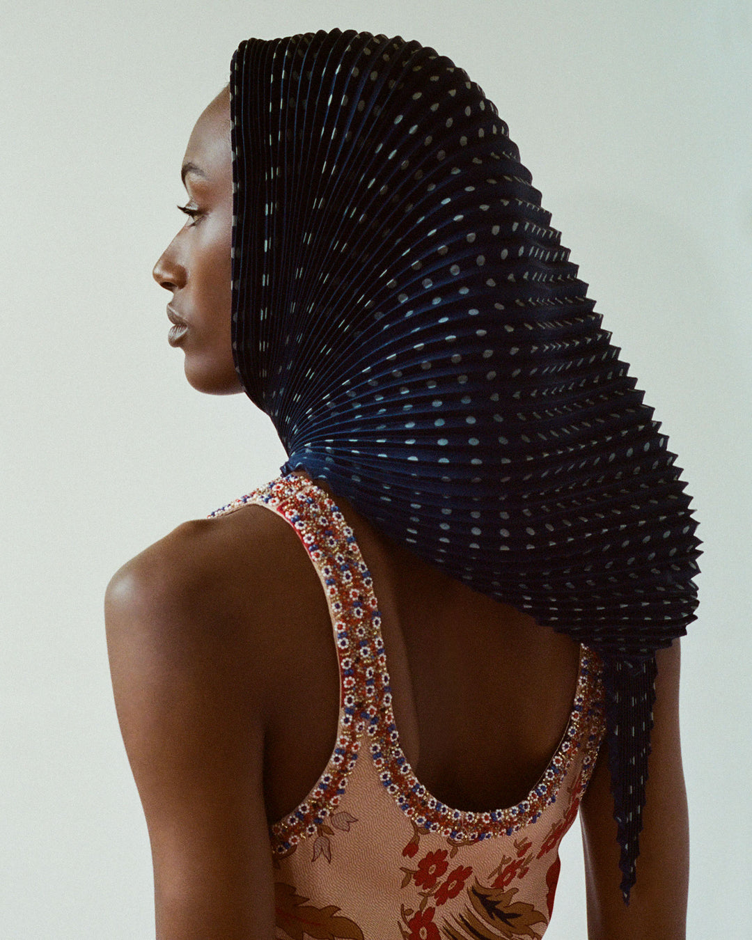 A person stands in profile, sporting a unique headpiece that fans out elegantly. They wear the Adele Vintage Poppy Dress, featuring a hand-beaded top with floral patterns and brightly embellished trim against a neutral background.