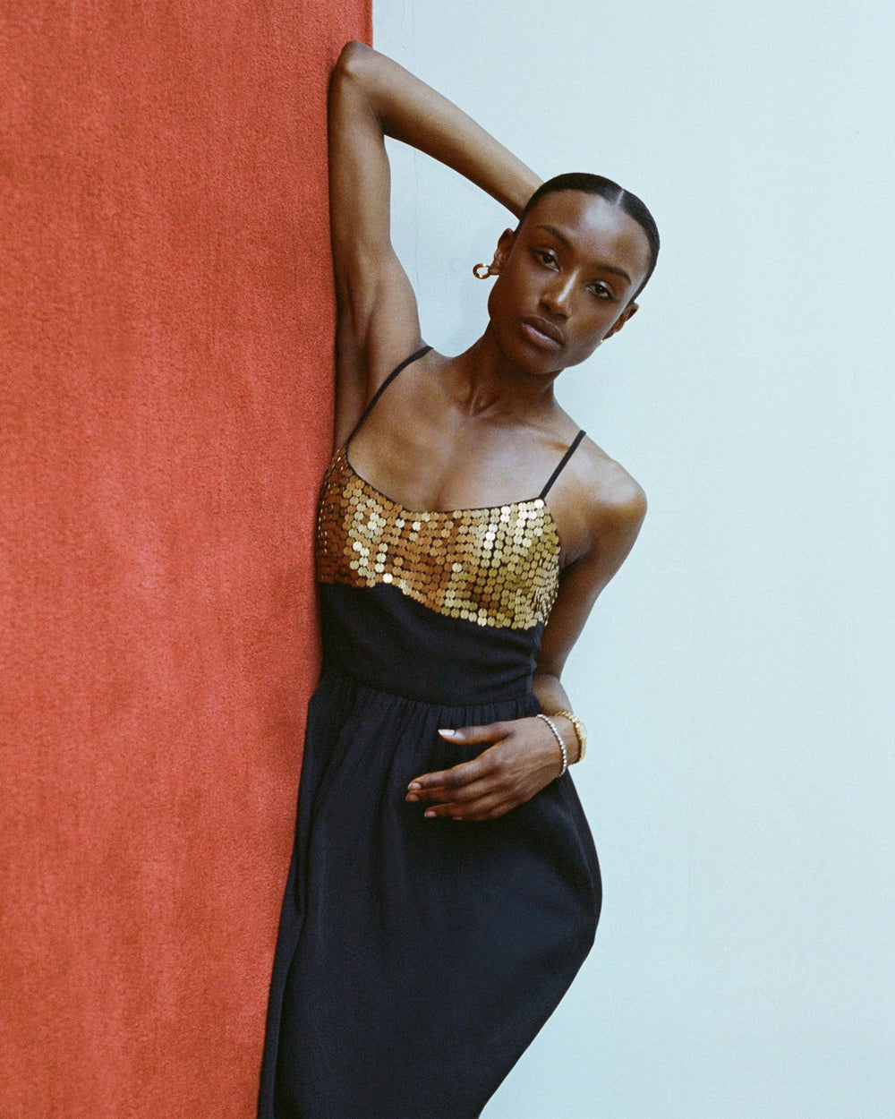 Wearing the Autumn Blackjack Coin Dress, a person elegantly poses against a red wall, with one arm resting above their head.